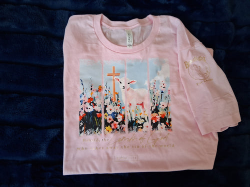 Light pink t-shirt with a religious design on a plush, dark blue background. Design consists of a lamb by a wooden cross, amidst wildflowers. Gold foil text below reads: "Behold, the Lamb of God who takes away the sins of the world. John 1:29"
