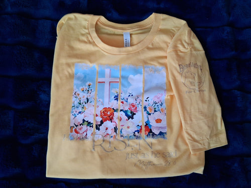 Yellow t-shirt on plushy, dark blue background. Design on shirt is of a white cross amid a bed of roses, in a painterly style. Gold foil text says, "He has risen, just as He said. Matthew 28:6"