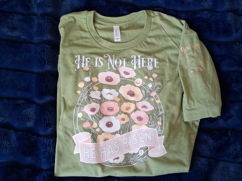Green t-shirt with floral design and "He is not here, He has risen!" text. Shirt is on a dark blue plush background.