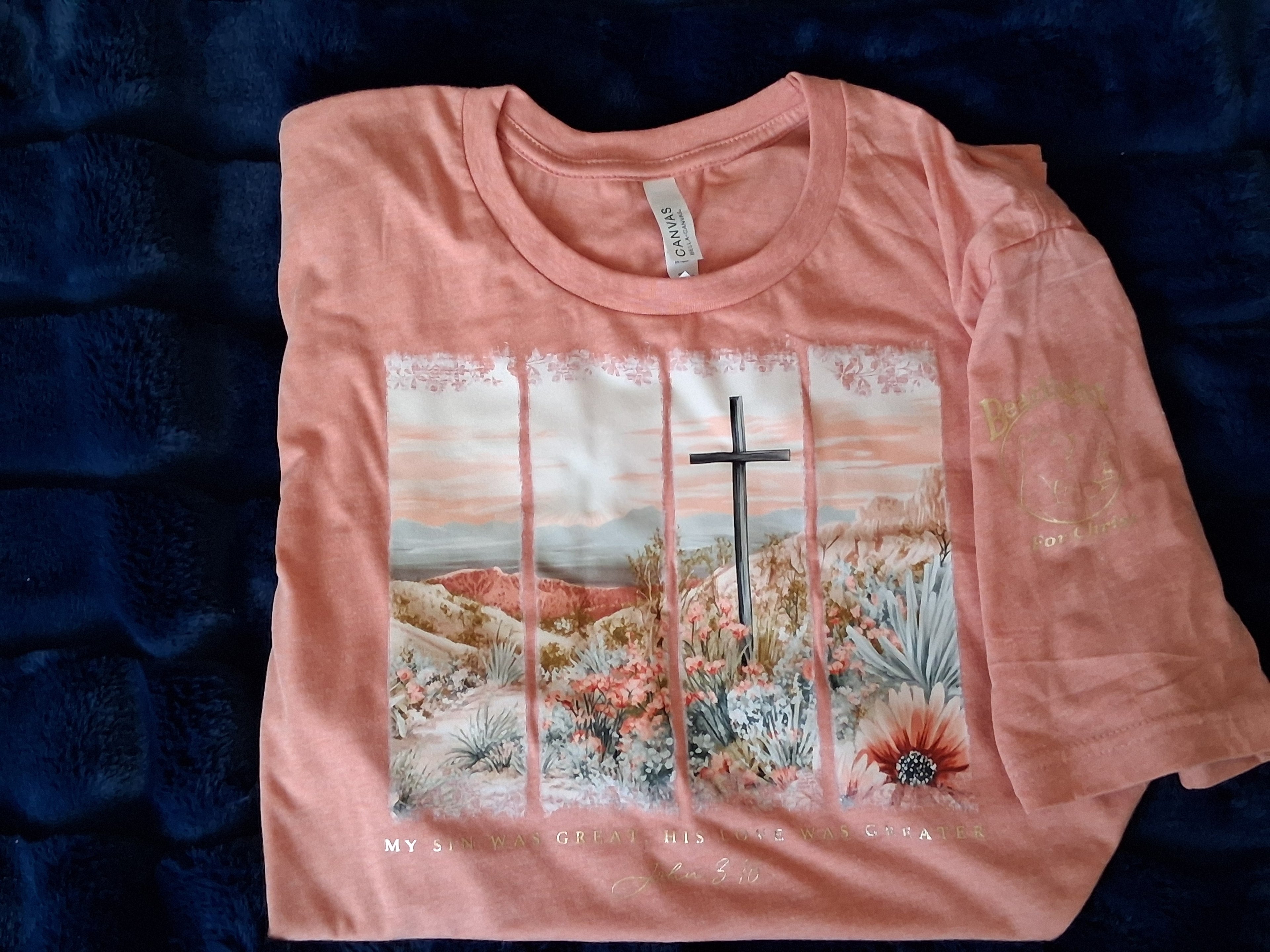 light orange t-shirt with a wooden cross in a desert-scape in spearmint and orange tones. Gold foil text says, "My sin was great, His love was greater. John 3:15". Shirt is on a plushy, dark blue background.
